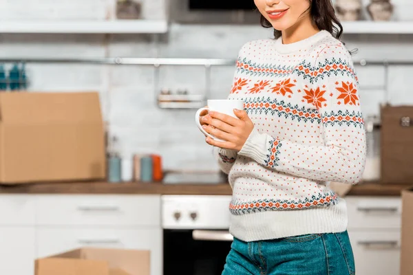 Partial view of young woman holding cup of coffee in kitchen with cardboard boxes during relocation in new home — Stock Photo