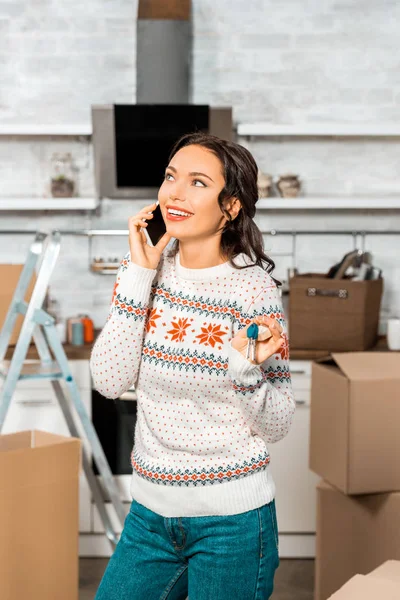 Joven mujer sonriente hablando en el teléfono inteligente y sosteniendo las llaves en la cocina en el nuevo hogar - foto de stock