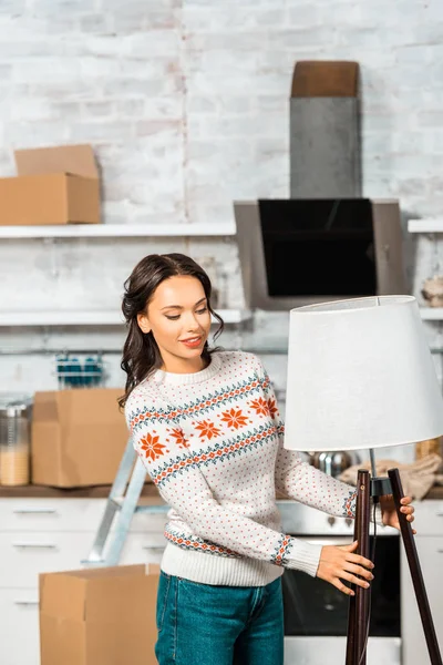 Mujer atractiva feliz poner la lámpara en la cocina durante la reubicación en un nuevo hogar - foto de stock