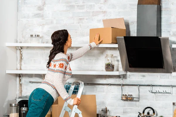 Visão traseira da mulher colocando caixa de cartão na prateleira na cozinha durante a realocação na nova casa — Fotografia de Stock