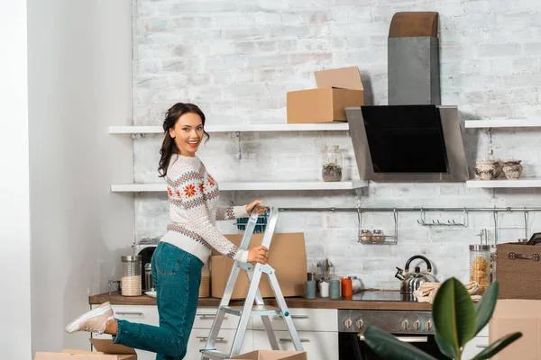 Смеющаяся молодая женщина, стоящая на лестнице на кухне с картонными коробками во время переезда в новый дом — стоковое фото