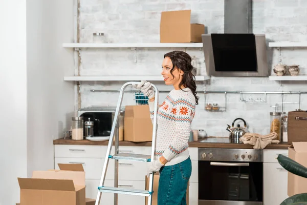 Vista lateral de la joven en guantes de trabajo que llevan escalera en la cocina durante la reubicación en el nuevo hogar - foto de stock
