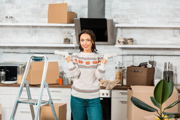 Beautiful young woman in working gloves doing shrug gesture near ladder in kitchen during relocation in new home — Stock Photo