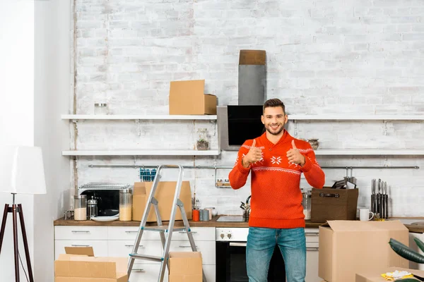 Heureux jeune homme fait pouces vers le haut dans la cuisine avec des boîtes en carton lors de la réinstallation dans la nouvelle maison — Photo de stock