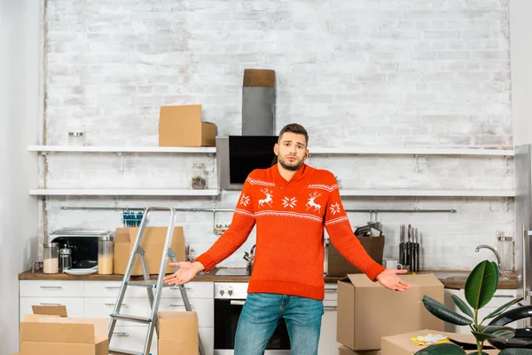 Young man doing shrug gesture in kitchen with cardboard boxes during relocation in new home — Stock Photo