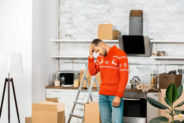 Upset young man holding forehead in kitchen with cardboard boxes during relocation in new home — Stock Photo