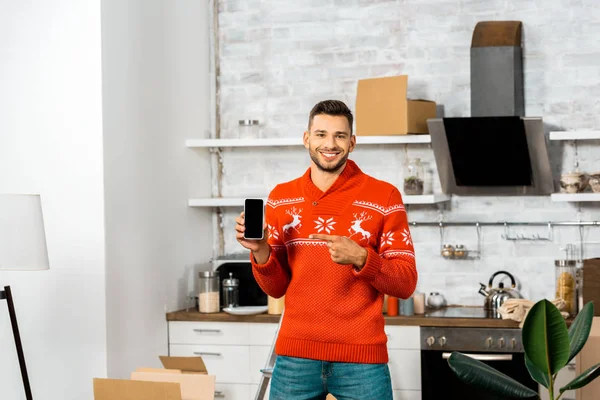 Happy young man pointing by finger at smartphone with blank screen in kitchen with cardboard boxes during relocation in new home — Stock Photo