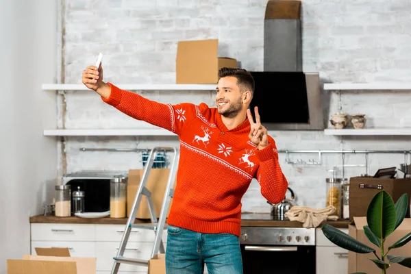 Счастливый молодой человек делает знак мира и делает селфи на смартфоне на кухне с картонными коробками во время переезда в новый дом — стоковое фото