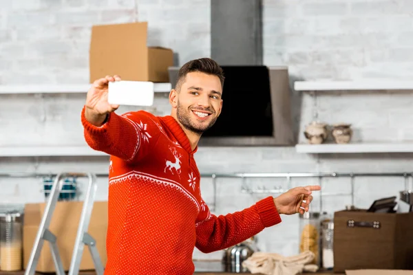 Lächelnder Mann macht Selfie auf Smartphone und zeigt mit dem Finger in Küche mit Pappkartons beim Umzug ins neue Zuhause — Stockfoto