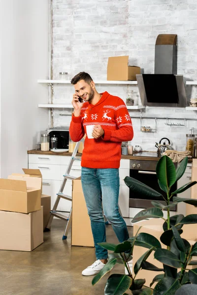 Улыбающийся мужчина с чашкой кофе разговаривает на смартфоне на кухне с картонными коробками во время переезда в новый дом — стоковое фото