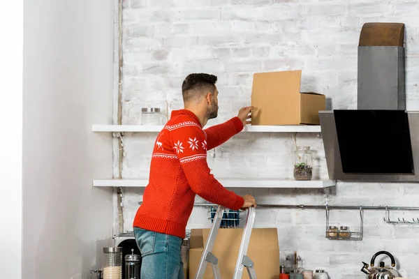 Vue arrière de l'homme debout sur l'échelle et mettre la boîte en carton sur l'étagère dans la cuisine lors de la réinstallation dans une nouvelle maison — Photo de stock