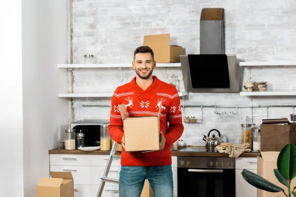 Homme souriant avec boîte en carton dans la cuisine avec boîtes en carton lors de la réinstallation dans une nouvelle maison — Photo de stock