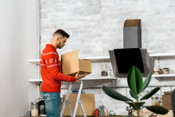 Vue latérale de l'homme debout sur l'échelle avec boîte en carton dans la cuisine lors de la réinstallation dans une nouvelle maison — Photo de stock