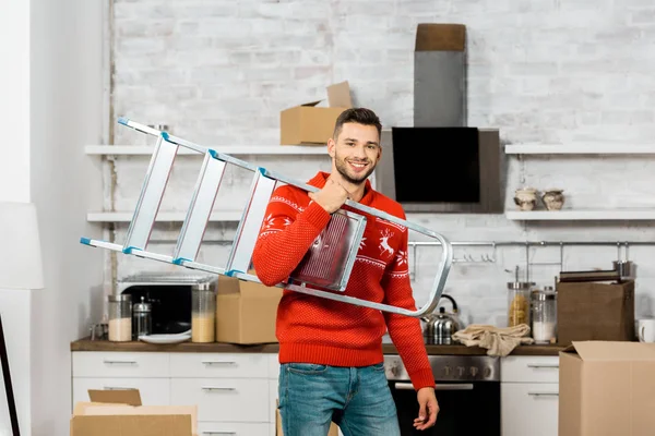 Cheerful young man carrying ladder in kitchen with cardboard boxes during relocation in new home — Stock Photo