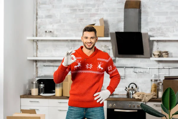 Happy young man in working gloves doing ok sign gesture in kitchen with cardboard boxes during relocation in new home — Stock Photo