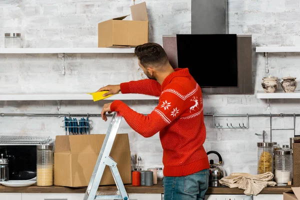 Vue arrière de l'homme debout sur l'échelle et essuyant la poussière des étagères lors du déménagement dans une nouvelle maison — Photo de stock