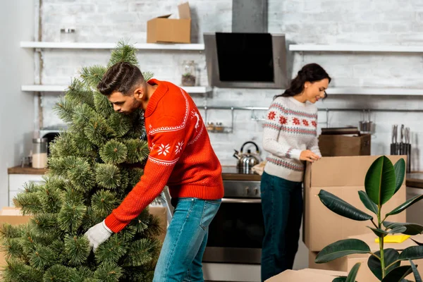 Young man putting christmas tree in kitchen while his girlfriend standing behind at home — Stock Photo