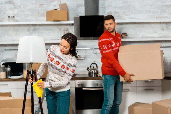 Attractive young woman wiping dust from lamp while her boyfriend carrying cardboard box in kitchen at new home — Stock Photo