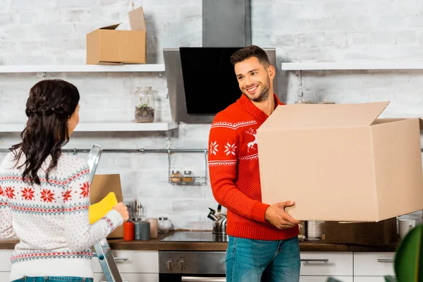 Cheerful man holding cardboard box and talking to girlfriend in kitchen at new home — Stock Photo