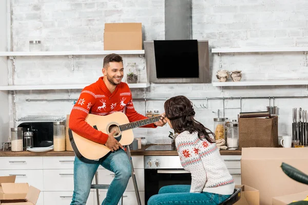 Сміється молодий чоловік грає на акустичній гітарі подрузі, сидячи на кухні нового будинку — стокове фото