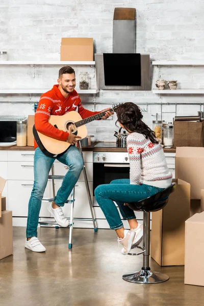 Улыбающийся мужчина, играющий на акустической гитаре с подругой, сидящей на кухне нового дома — стоковое фото