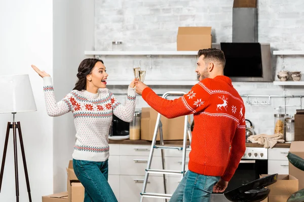 Animado jovem casal comemorando realocação em nova casa e clinking por copos de champanhe na cozinha — Fotografia de Stock
