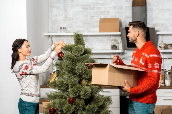 Улыбающаяся молодая пара украшает рождественскую елку безделушками на кухне дома — стоковое фото