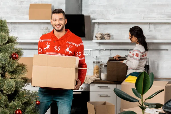 Mann hält Pappschachtel mit Christbaumkugeln zum Dekorieren des Weihnachtsbaums, während seine Freundin zu Hause in der Küche steht — Stockfoto