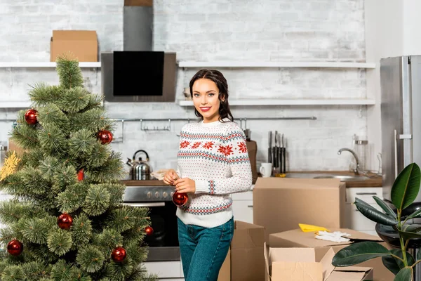 Beautiful young woman decorating christmas tree by baubles in kitchen at home — Stock Photo