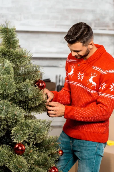 Glücklicher junger Mann schmückt Weihnachtsbaum mit Kugeln in der heimischen Küche — Stockfoto