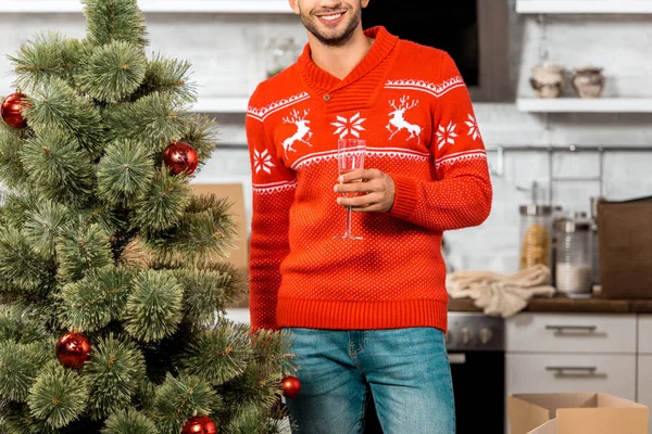 Частичный вид улыбающегося мужчины, празднующего с бокалом шампанского возле рождественской елки дома — стоковое фото