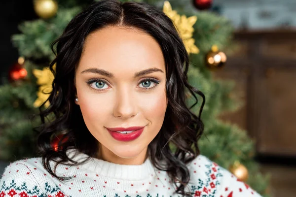 Retrato de mujer joven atractiva mirando la cámara cerca del árbol de Navidad en casa - foto de stock