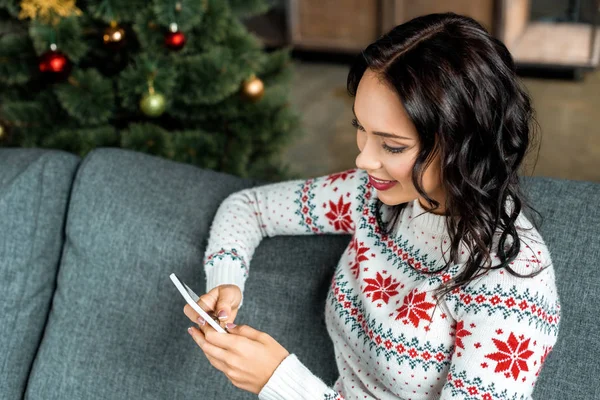 Mulher sorrindo usando smartphone no sofá perto da árvore de natal em casa — Fotografia de Stock