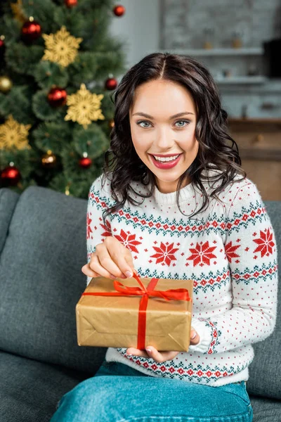 Усміхнена молода жінка з подарунковою коробкою на дивані біля ялинки вдома — стокове фото