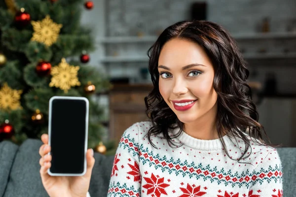 Schöne Frau zeigt Smartphone mit leerem Bildschirm auf Sofa in der Nähe des Weihnachtsbaums zu Hause — Stockfoto