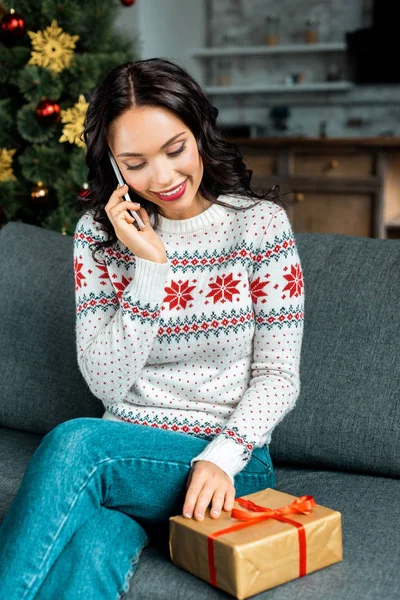 Junge Frau mit Geschenkbox spricht auf Smartphone auf Sofa in der Nähe des Weihnachtsbaums zu Hause — Stockfoto