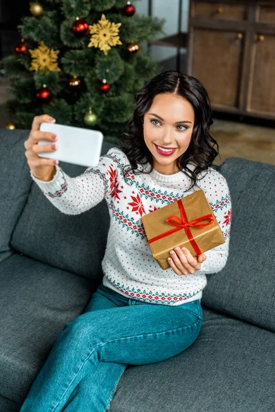 Усміхнена жінка бере селфі на смартфон з подарунковою коробкою на дивані біля ялинки вдома — стокове фото