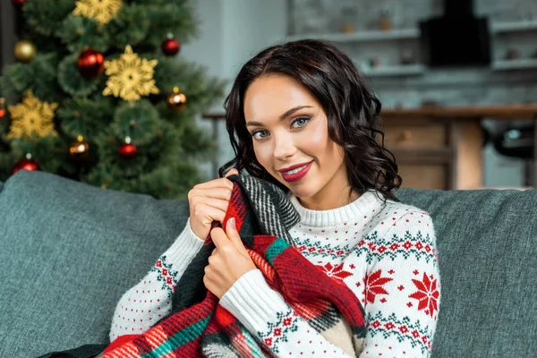 Retrato de mulher bonita sentada no sofá com cobertor perto da árvore de natal em casa — Fotografia de Stock