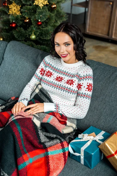 Красивая молодая женщина сидит под одеялом на диване с подарочными коробками возле рождественской елки дома — стоковое фото