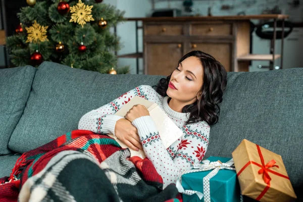 Bella giovane donna con libro dormire sul divano sotto coperta con regali vicino albero di Natale a casa — Foto stock