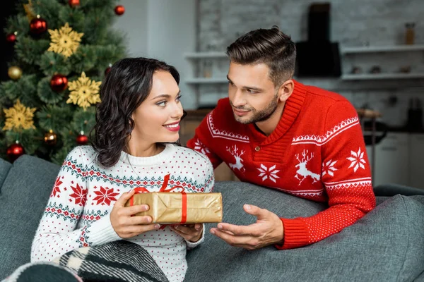 Smiling young man gifting present box to attractive girlfriend on sofa near christmas tree at home — Stock Photo