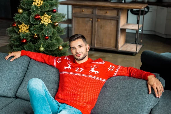 Junger schöner Mann sitzt auf Sofa in der Nähe von Weihnachtsbaum zu Hause — Stockfoto