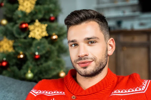 Retrato de un hombre guapo mirando a Camer y sentado en el sofá cerca del árbol de Navidad en casa — Stock Photo