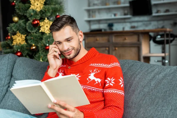 Jovem bonito ler livro e falando no smartphone perto da árvore de natal em casa — Fotografia de Stock