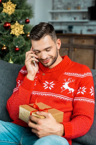 Uomo sorridente che parla su smartphone e tiene la scatola regalo sul divano vicino all'albero di Natale a casa — Foto stock