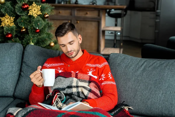 Junger Mann sitzt auf Sofa unter Decke und liest Buch und hält Tasse Kaffee in der Nähe von Weihnachtsbaum zu Hause — Stockfoto