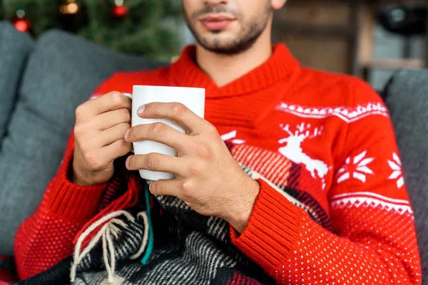 Vista parcial del hombre sentado en el sofá debajo de la manta sosteniendo la taza de café cerca del árbol de Navidad en casa — Stock Photo