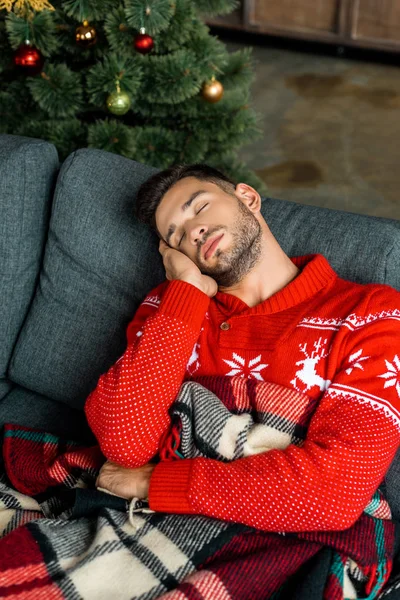 Schöner junger Mann schläft auf Sofa unter Decke in der Nähe von Weihnachtsbaum zu Hause — Stockfoto