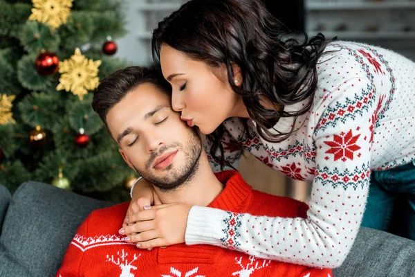 Селективное внимание привлекательной женщины целующей спящего бойфренда на диване возле рождественской елки дома — стоковое фото