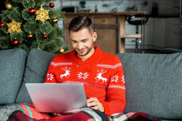 Fröhlicher Mann sitzt auf Sofa unter Decke und benutzt Laptop in der Nähe von Weihnachtsbaum zu Hause — Stockfoto